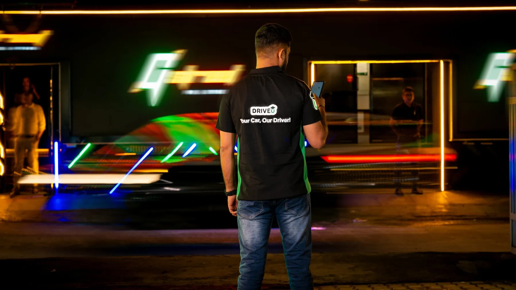 A DriveU driver stands still while a car streaks past in a long exposure, his reflective logo sharp against the light trails